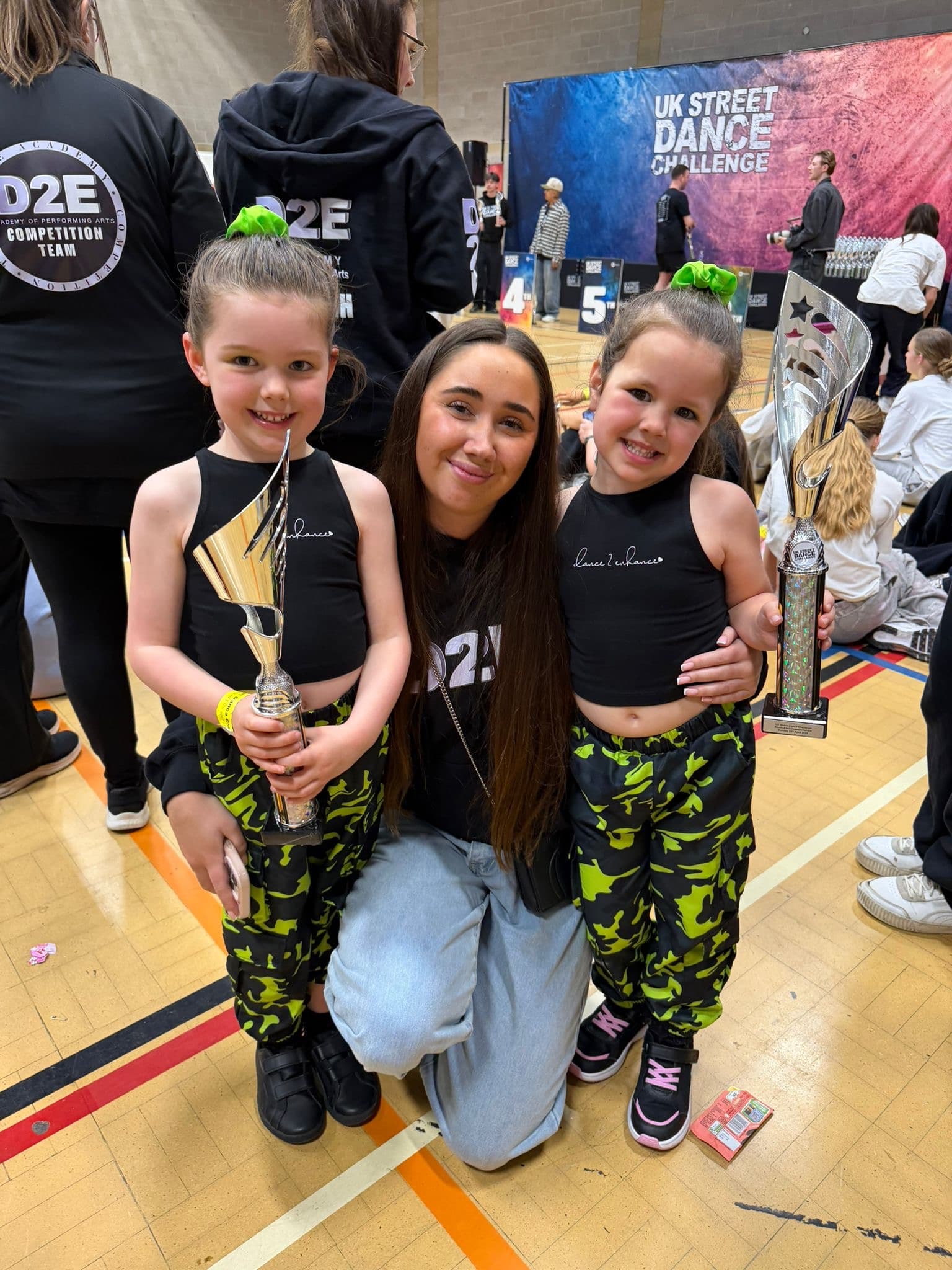 Two D2E dancers in neon camo holding trophies with their teacher at the UK Street Dance Challenge